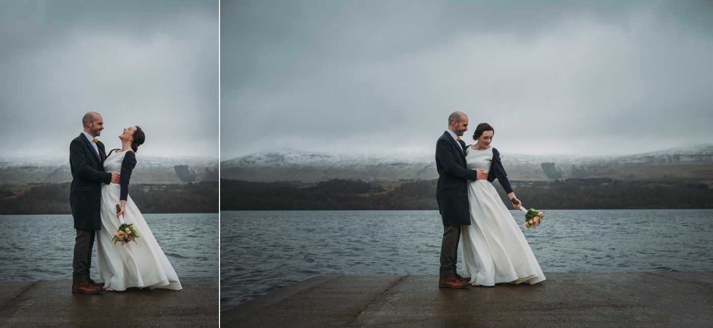 A newly married couple laugh and hug at Ardenoaig, Loch Tay and mountains in the background on their scottish elopement.