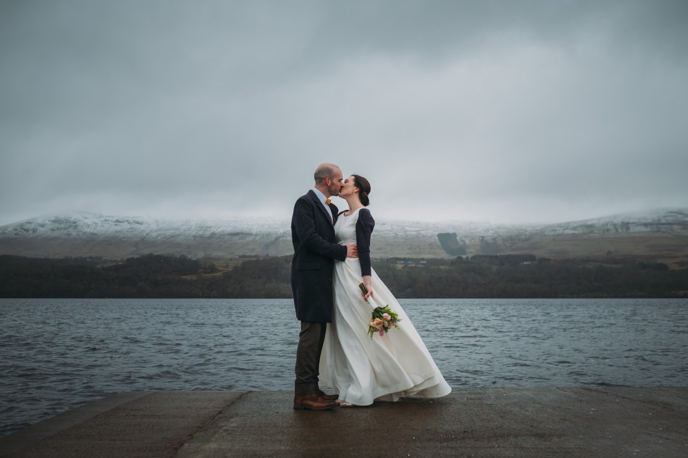 A newly married couple kiss at Ardenoaig, Loch Tay and mountains in the background on their scottish elopement.