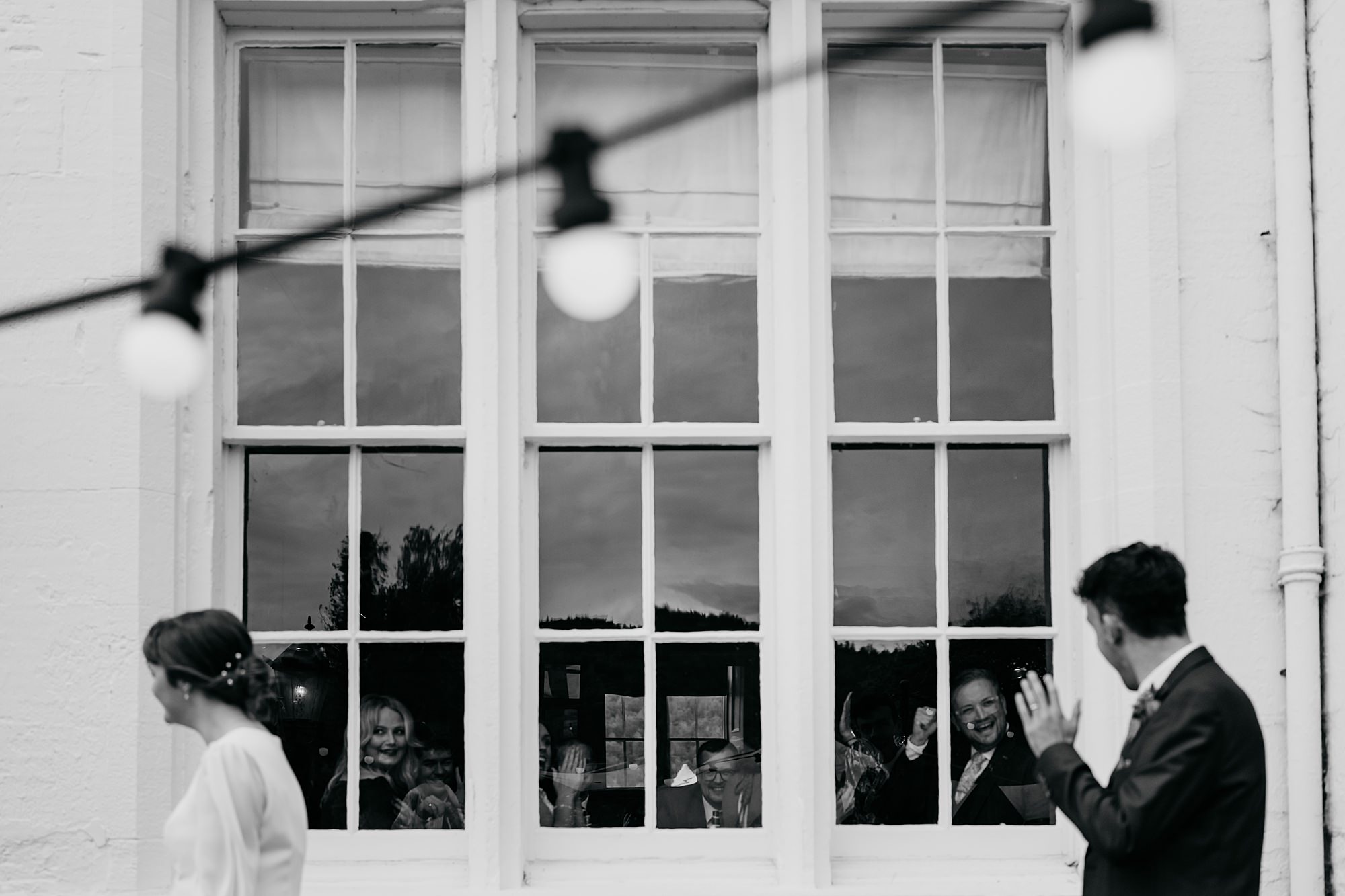 bride and groom walk past pub window. Their guests wave at them and they wave back. relaxed wedding photography for a Hermitage elopement.
