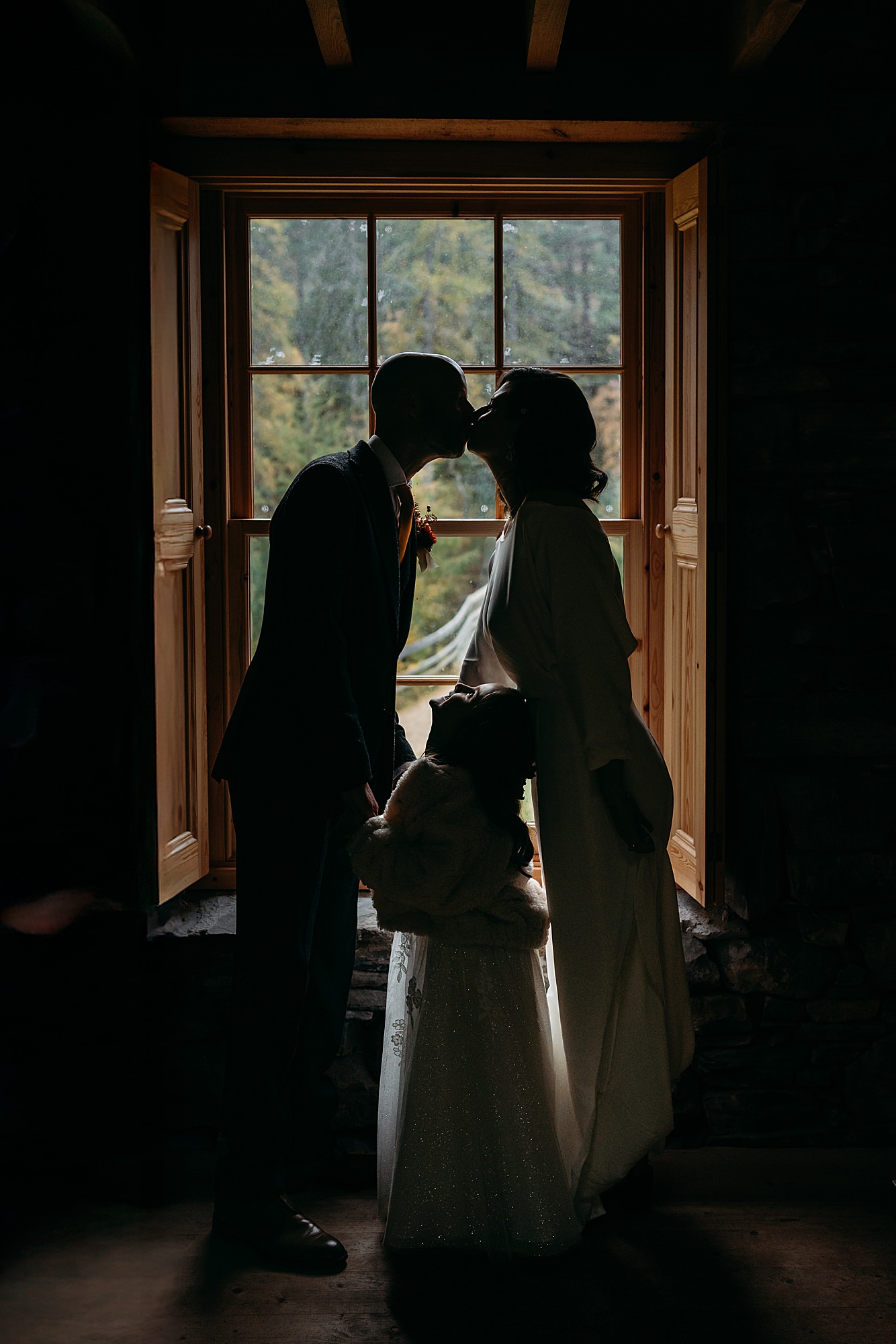 A couple kiss as their daughter looks on, they are all silhouette in the window. Relaxed wedding photography in Braemar with Coorie Wedding Planning.