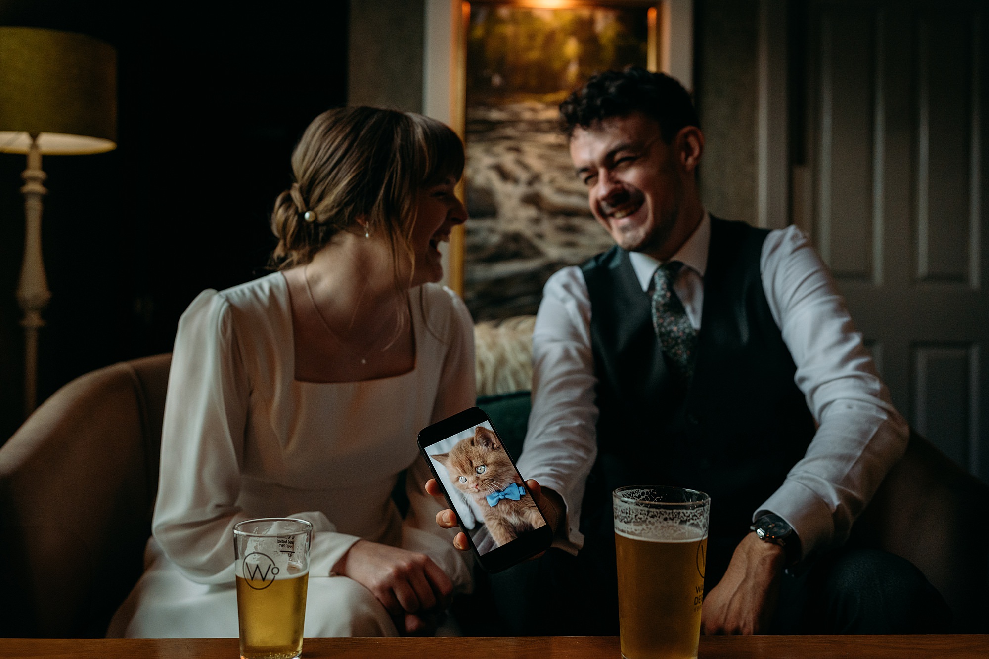 Relaxed wedding photograph of newlyweds laughing over a photo of a cat on the grooms phone. 