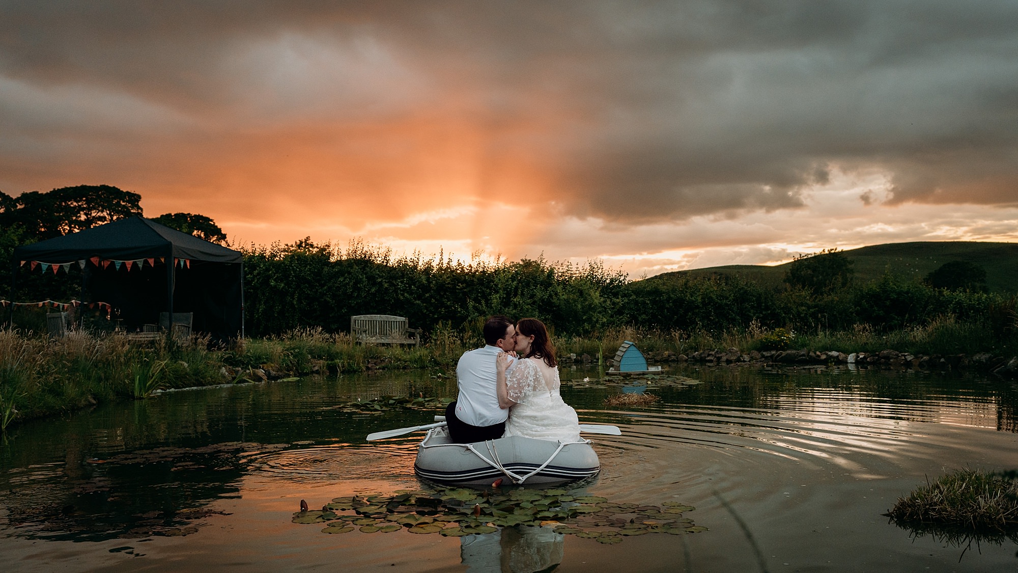 A couple kiss at sunset on a small dingy in a big pond at their garden wedding. Relaxed wedding photography in the Scottish Borders.