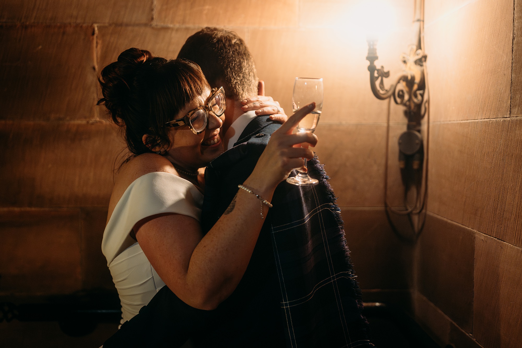 A bride smiles as she is hugged closely by her husband. She hold a champagne glass. Relaxed wedding photography at Coats Paisley.