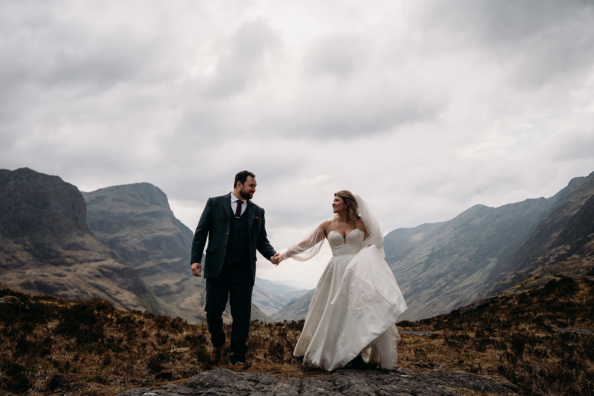 American newlyweds elope to Glencoe. They walk hand in hand with The Three Sisters mountain range behind them. relaxed wedding photography