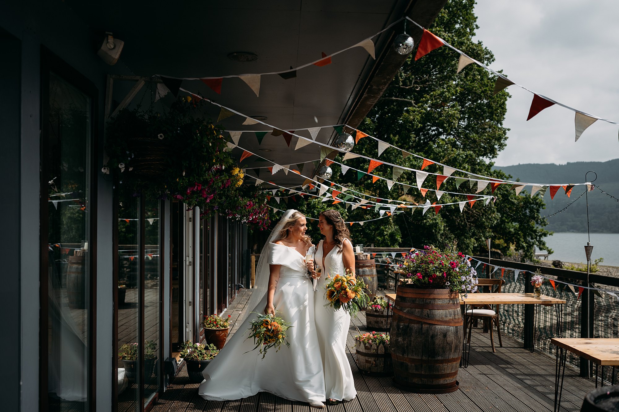 Two brides laugh together under colourful bunting at their Venachar lochside wedding. relaxed, inclusive wedding photography
