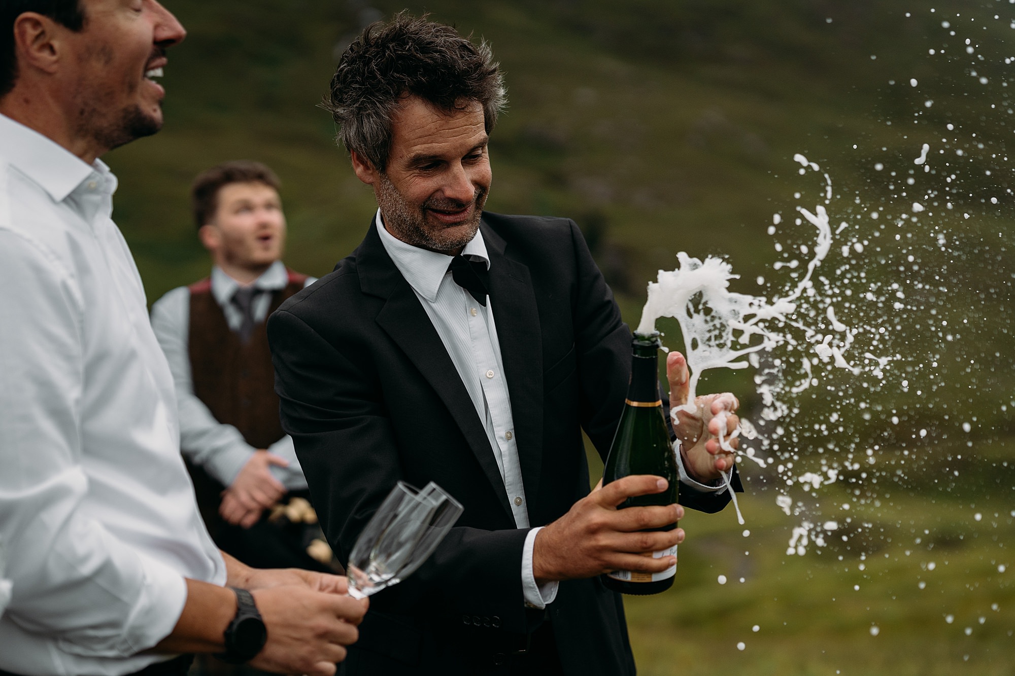 Man pops bottle of champagne in Glencoe, the cork goes flying and champagne spray everywhere. Grant MacLeod Scottish bagpiper looks surprised in background. relaxed wedding photography with French Wedding in Scotland