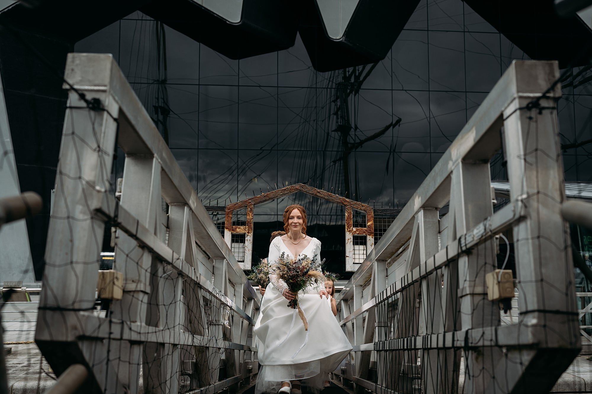 Bride boards Tall Ships Glasgow before her ceremony. Riverside Museum rises behind her. relaxed wedding photography in Glasgow.