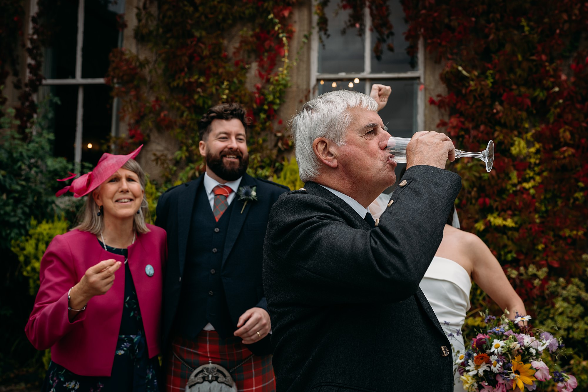 Brides Dad walks past downing champagne glass as family look on laughing. relaxed wedding photography in Loch Lomond.