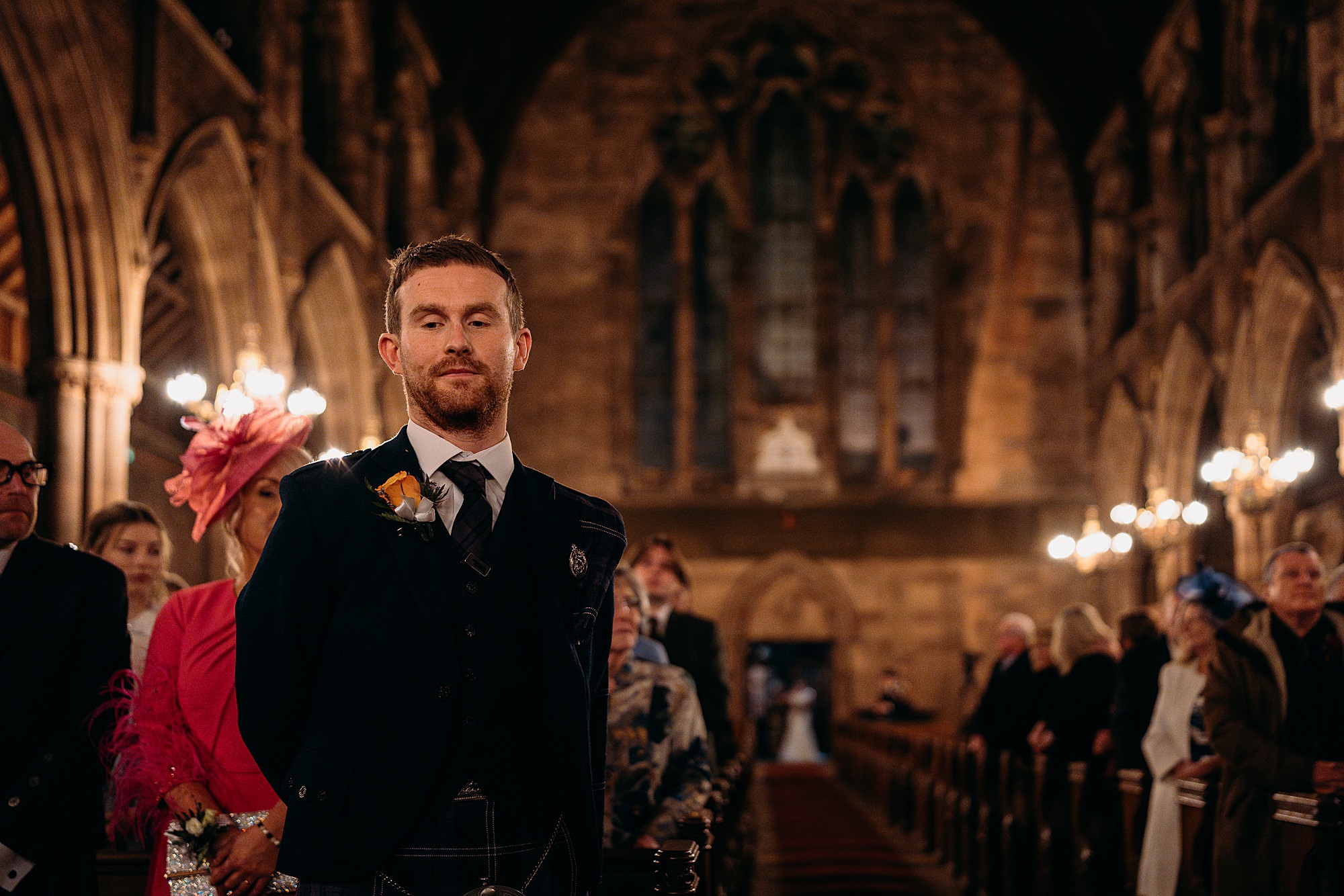 Groom waits for his bride at their Coats Paisley Twilight Wedding. She is at the end of the aisle, out of focus